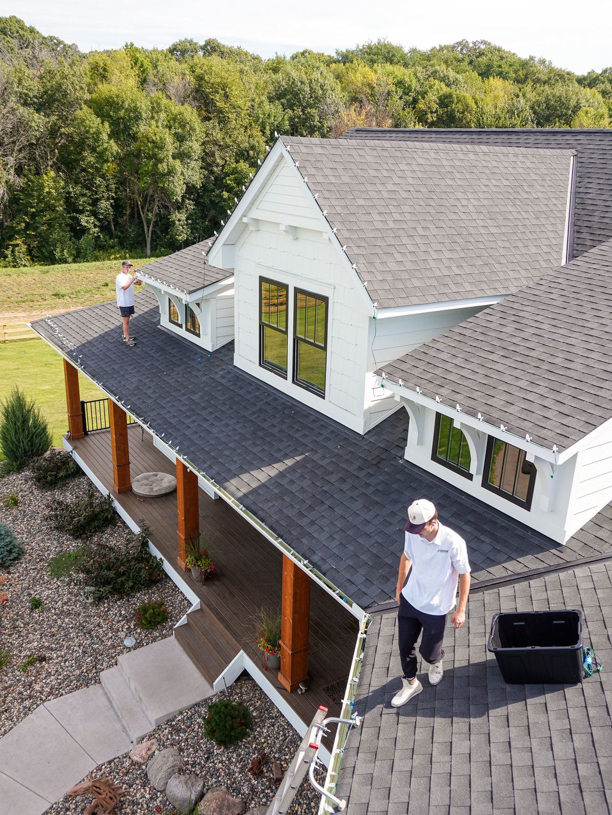 Two technicians stringing holiday lights across a modern farmhouse roof, seen from above