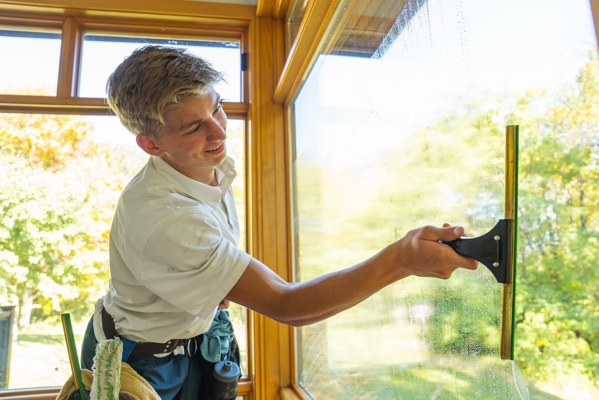 A smiling technician squeegeeing the inside of a sunroom window, fall trees outside