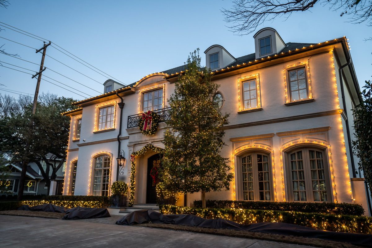 A white two-story home at dusk with warm white lights outlining the roofline and every window, lit garland over the front door and lights in the hedges