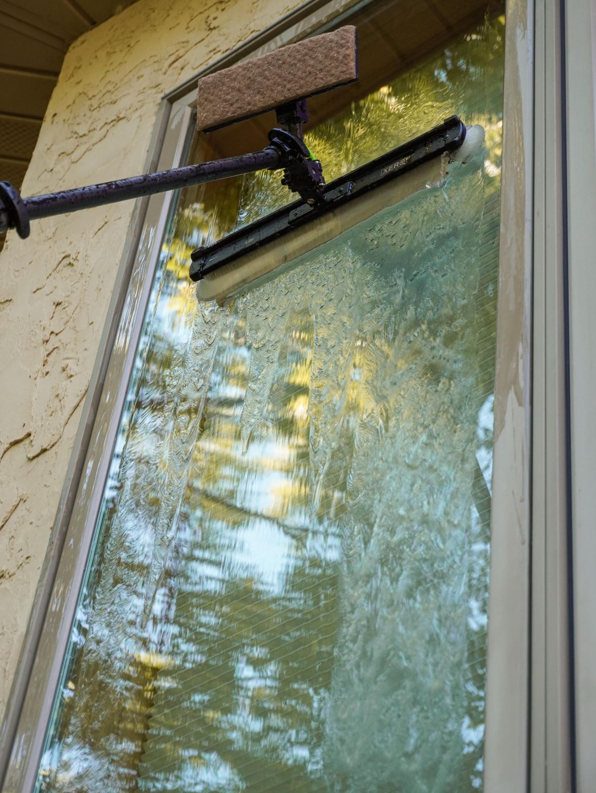 Water-fed pole brush scrubbing an upper pane, rinse water sheeting down the glass
