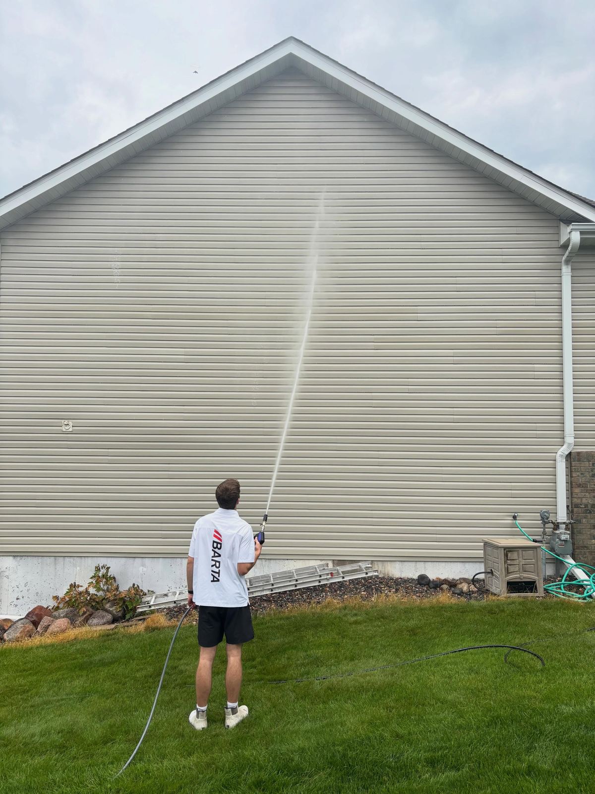 Technician rinsing a tall vinyl-sided gable wall from the back lawn