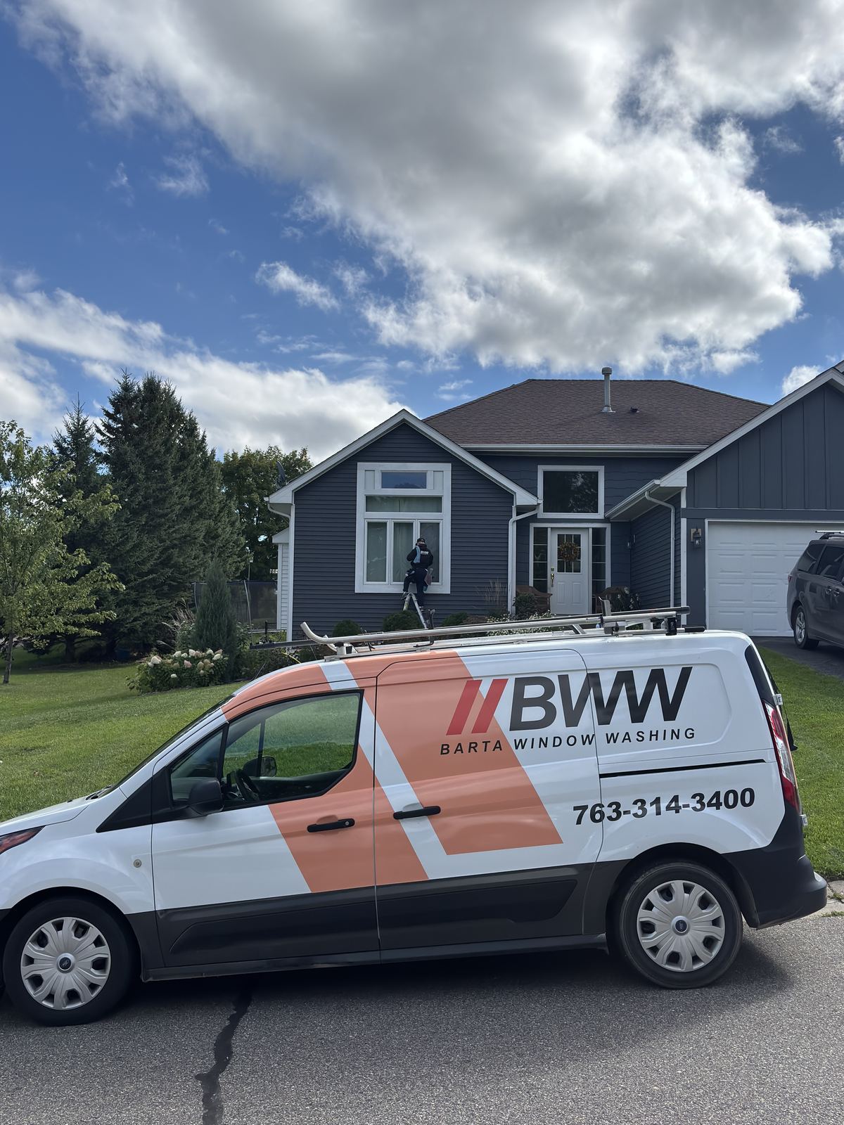 Barta technician on a ladder cleaning the front windows of a Buffalo home, with the Barta van out front