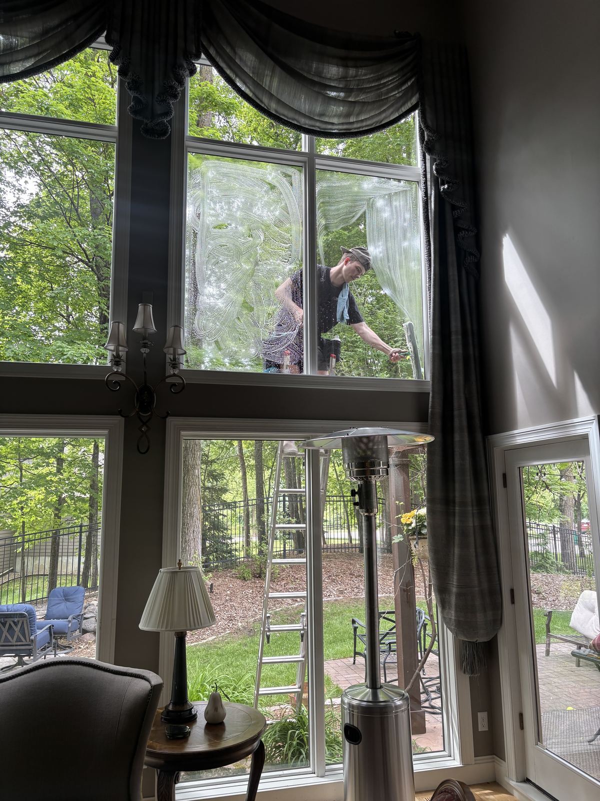 Seen from inside, a Barta crew member on a ladder cleans the upper panes of a two-story wall of windows in a Minnetrista home
