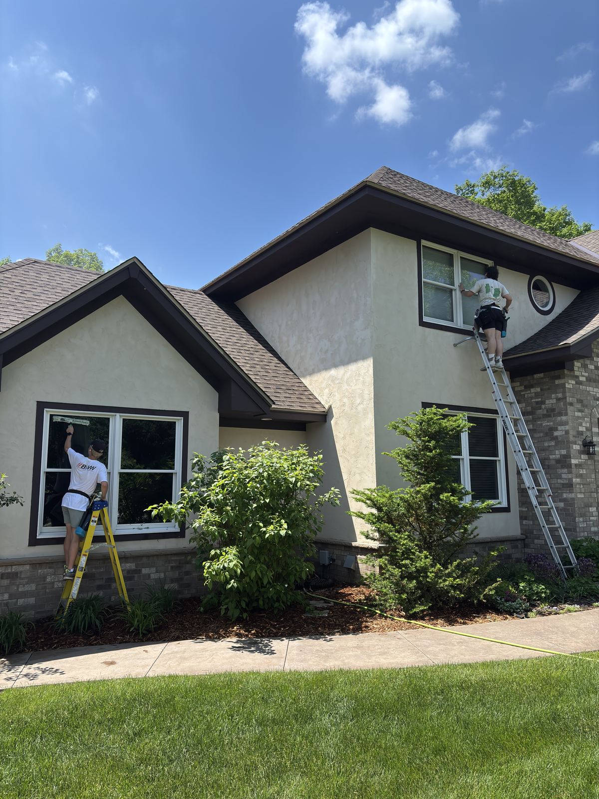 Two Barta crew members cleaning the front windows of a stucco home in Mound, one on an extension ladder and one on a stepladder