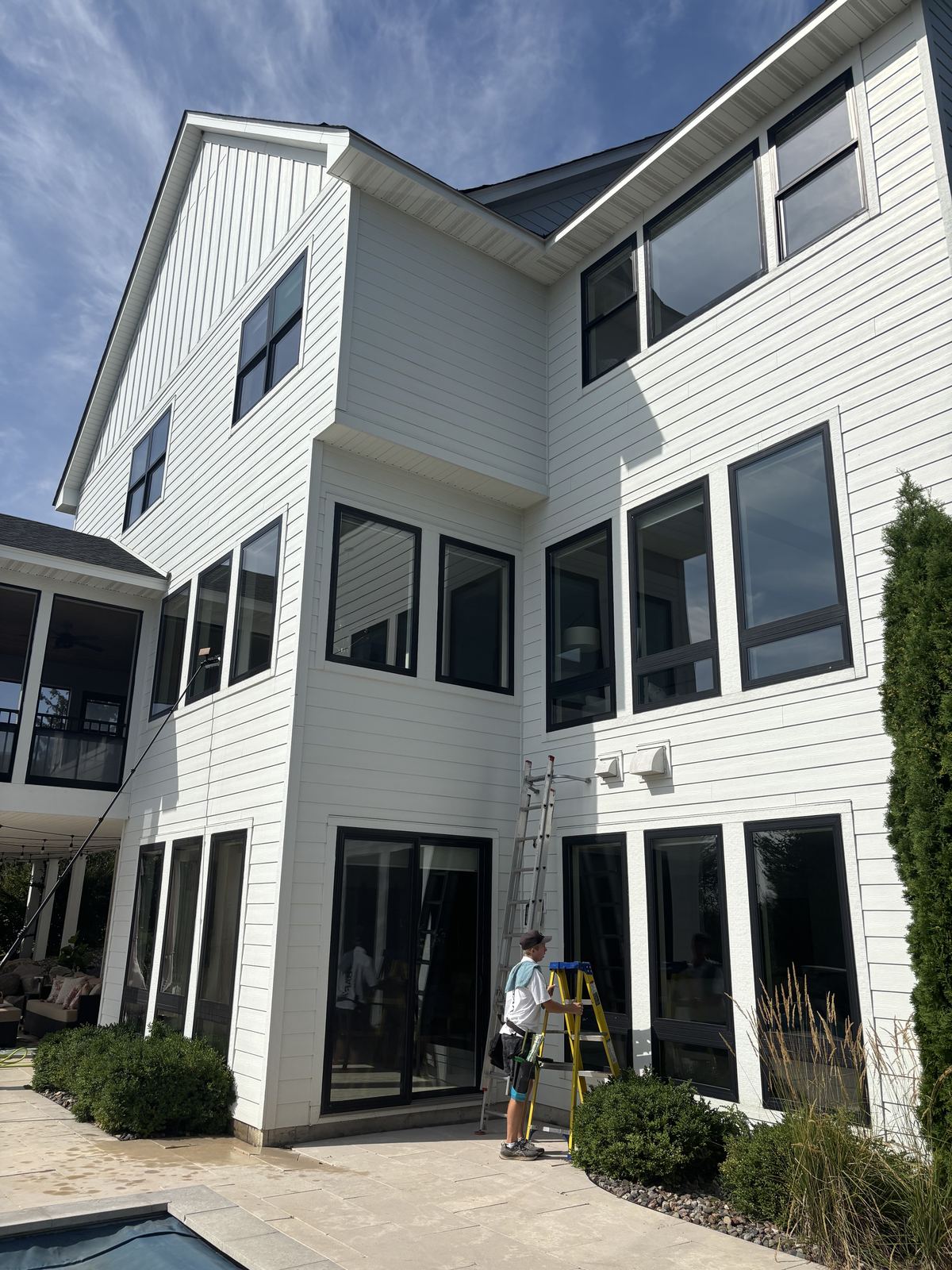 Barta technician cleaning the back windows of a three-story Orono home with a water-fed pole and a ladder