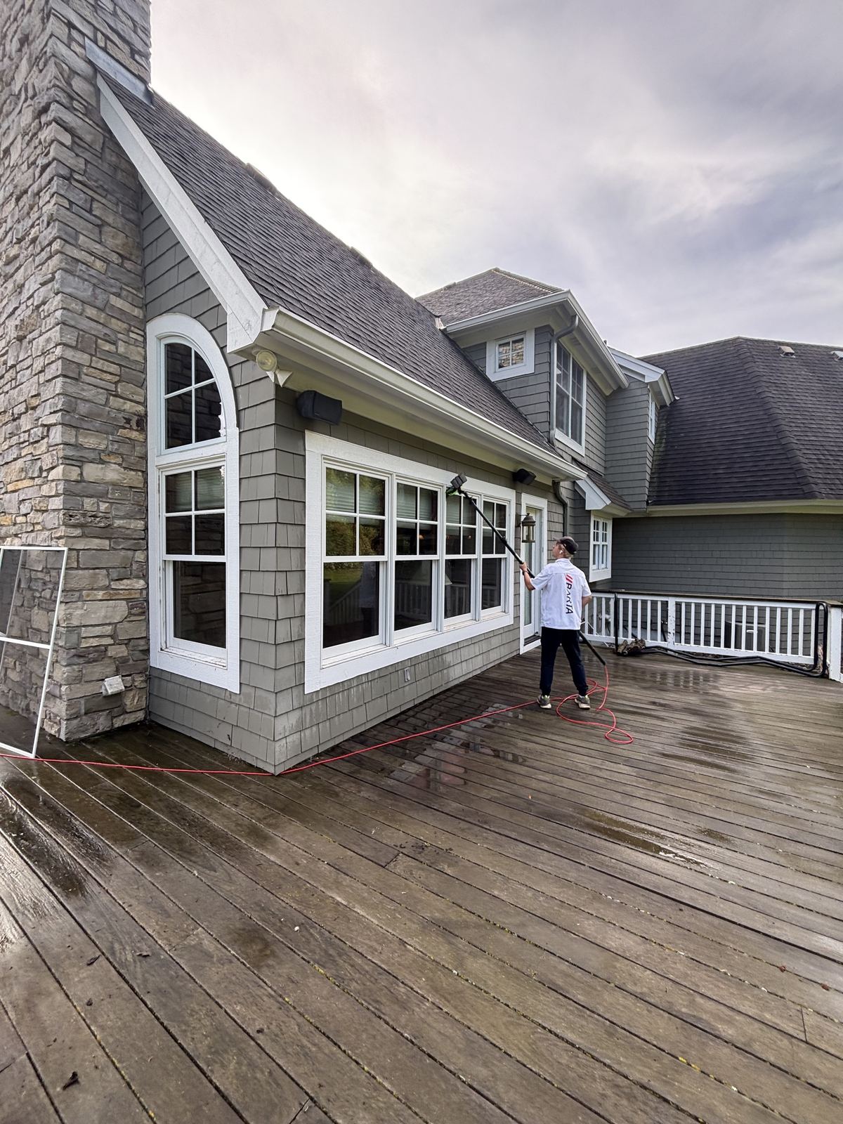 Crew member cleaning a row of arched and square windows along a wet deck with a water-fed pole, beside a stone chimney