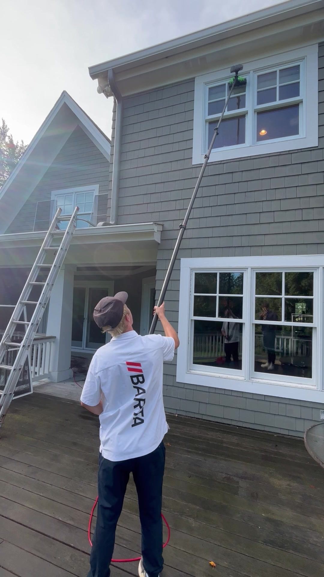 Barta crew member on a deck guiding a water-fed pole up to a second-story window of a gray shingle-sided home near Watertown