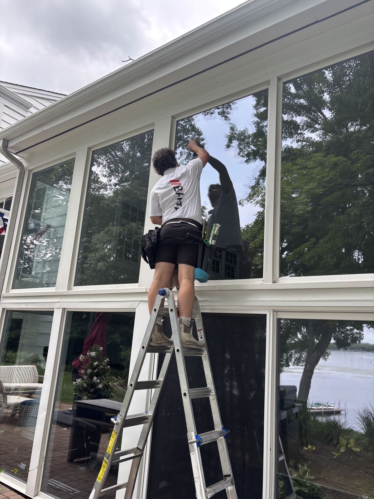 Barta Window Washing technician on a ladder cleaning the windows of a lake-facing sunroom at a Wayzata home
