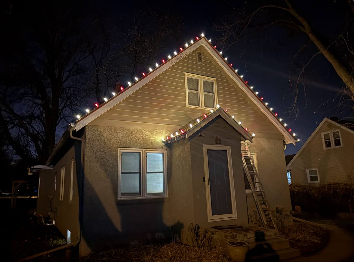 Red and white holiday lights installed along a bungalow's roofline at night