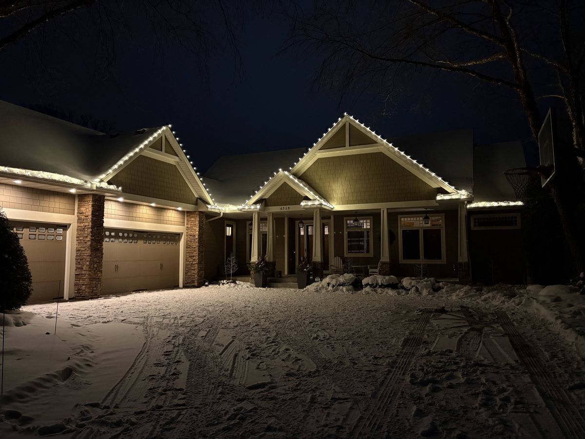 White holiday lights tracing the gables of a craftsman-style home above a snow-covered driveway
